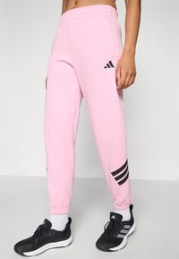 Lyserøde sweatpants med elastisk talje og afsluttede ankler. Har sort Adidas-logo og tre sorte striber på venstre ben.
