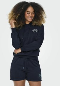 Completo con felpa blu navy e pantaloni corti realizzato in tessuto morbido, con cappuccio regolabile e dettaglio del logo frontale. Texture liscia, vestibilità comoda.