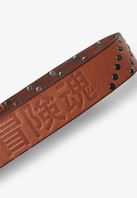 Brazalete de cuero marrón con caracteres en relieve y tachuelas de metal negro a lo largo del borde. La textura es suave con un acabado mate.