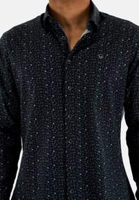Chemise à manches longues avec boutons de couleur navy foncé, ornée d'un petit motif floral bleu clair. Elle présente un col ouvert et des boutons noirs. Texture de tissu douce.