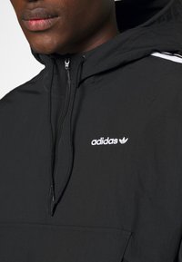 Černá mikina s polovičním zipem značky Adidas, se stažitelnou kapucí a bílým logem. Hladká tkanina s bočními pruhy a klokankovou kapsou.