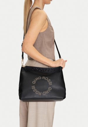 Mujer con conjunto beige lleva un gran bolso de hombro negro de Gino Rossi con logo circular en tachuelas doradas y correa con nombre de la marca en relieve.