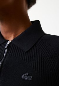 Černé pletené polo s žebrovanou strukturou, zipovým zapínáním a plochým límcem. Na hrudi má texturované logo, které nabízí elegantní design.