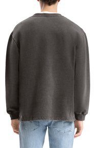 Uomo che indossa una felpa grigia scura a maniche lunghe e jeans azzurri chiari, ripreso da dietro su uno sfondo bianco.