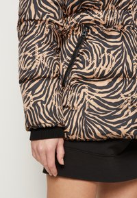 Doudoune beige et noire imprimé zèbre avec poche zippée diagonale, manches resserrées, portée sur une jupe noire, montrant une main et une cuisse.