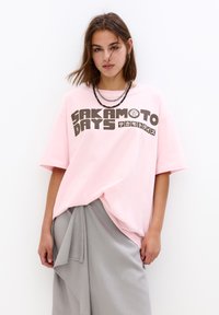 Lyserød bomulds oversized t-shirt med fed sort tekst "SAKAMOTO DAYS" og japanske tegn. Løs pasform, rund halsudskæring, korte ærmer.