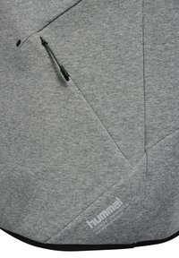 Gråt tekstureret stof med diagonale sømme, sort lynlås træk, og hvid hummel-logo med tekst nær den nederste kant.