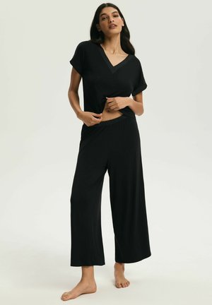 Kobieta stojąca boso, ubrana w czarną bluzkę z krótkim rękawem i dekoltem w serek oraz luźne czarne spodnie o szerokich nogawkach na neutralnym tle.