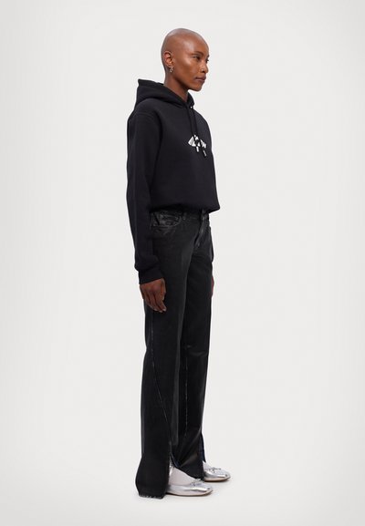 Black hooded sweatshirt with a front graphic, paired with loose black jeans featuring side slits, and silver shoes.