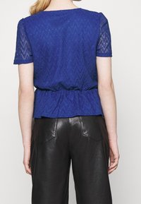 Žena v královsky modrém krátkém rukávu krajkovém topu se zigzagovým vzorem a černých kožených kalhotách, stojící s odvráceným pohledem od kamery.