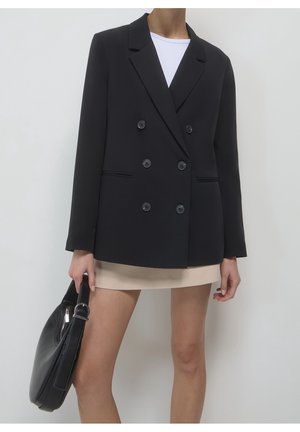 Frau trägt einen schwarzen, zweireihigen Blazer, einen beigen Rock, ein weißes Top und hält eine schwarze Handtasche an ihrer Seite.