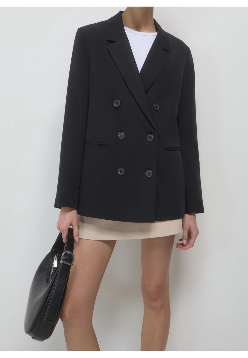 Femme portant un blazer noir croisé, une jupe beige, un haut blanc, et tenant un sac à main noir à ses côtés.