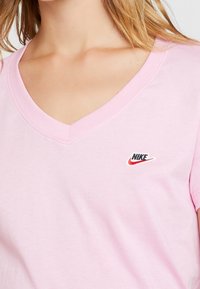 Rosa bomullströja med v-ringning. Har en liten broderad svart och röd Nike-logotyp på bröstet. Mjuk textur, korta ärmar.