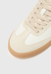 Beige athletic shoe featuring a suede and leather upper, round toe, textured laces, and a gum rubber sole for contrast.