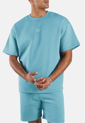 Mann trägt hellblaues Reebok-Kurzarm-Sweatshirt und passende Shorts, mit Ringen und Armband, vor einem schlichten weißen Hintergrund.