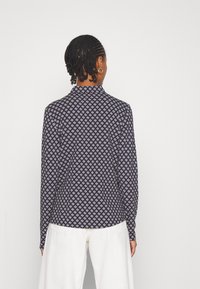 Femme aux cheveux tressés portant une chemise à manches longues à motifs bleu marine et un pantalon large blanc, de dos par rapport à l'appareil photo.