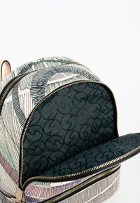 Mochila con un exterior de tela estampada en blanco roto y verde, con cremalleras doradas y un interior forrado en verde oscuro con un patrón de logo.
