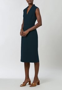 Femme portant une robe bleu marine sans manches, longueur genou, avec un col en V, associée à des sandales à talons carrés ouvertes de couleur beige, debout devant un fond uni.