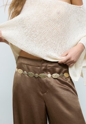 Persona che indossa pantaloni di raso marrone e un top lavorato a maglia color crema, mostrando una cintura decorativa in oro martellato intorno alla vita.