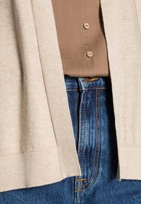 Beige cardigan made of soft knit fabric, featuring a relaxed fit, open front, and ribbed hem, paired with a brown button-up shirt and blue jeans.