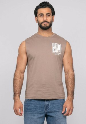 Homme barbu portant un débardeur marron clair avec un motif blanc et un jean bleu, debout devant un fond uni.