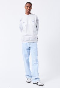 Sudadera gris con gráficos en blanco y azul, combinada con jeans de un azul claro. El modelo está de pie sobre un fondo sencillo, vistiendo zapatillas negras y blancas.