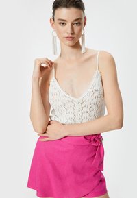 Top de encaje blanco con tirantes finos y escote en V, combinado con una falda envolvente rosa con un nudo lateral y detalles en dorado.