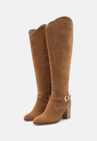 Καφέ σουέτ γόβες μέχρι το γόνατο με τετράγωνη γάμπα, που διαθέτουν διακοσμητική ζώνη και πόρπη, στρογγυλή μύτη και μαλακή υφή.