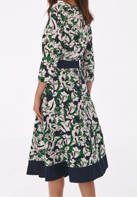 Abito floreale in navy, verde e bianco. Presenta maniche lunghe, una vita sottolineata e una gonna svasata con un orlo navy. Texture del tessuto liscia.