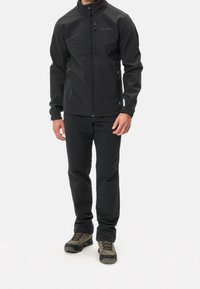 Veste softshell noire avec col montant, fermeture éclair à l'avant et panneau contrastant aux épaules ; portée avec un pantalon noir et des chaussures de randonnée.
