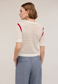Suéter blanco de punto con acentos rojos, de manga corta, con patrón calado, combinado con pantalones azules texturizados, visto desde la espalda.