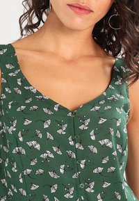 Top verde sin mangas con cuello en V, con un estampado de hoja de ginkgo blanca y detalles de botones en la parte delantera. Tejido liso y ligero.