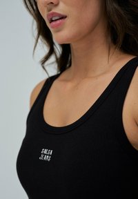 Top de tirantes negro con textura acanalada y escote redondo. Presenta un pequeño logo en relieve "Salsa Jeans" en la parte frontal. Textura suave, diseño ajustado.