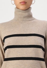 Beige garbó pulóver fekete vízszintes csíkokkal. Az anyag puhanak tűnik, bordás textúrával. Fitted gallérral rendelkezik.