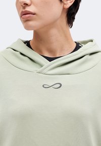 Personne aux cheveux courts portant un sweat à capuche vert clair avec un petit symbole noir d'infini sur la poitrine.