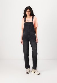 Zwarte denim overalls met verstelbare bandjes, twee voorzakken en een zijzak, gecombineerd met een lichtroze graphic t-shirt en sneakers.