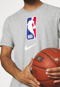Camiseta de algodón gris con el logo de la NBA en color y el swoosh blanco de Nike. Persona sostiene un balón de baloncesto marrón con la marca "Elite".