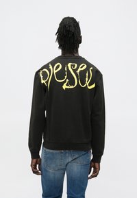 Μαύρο φούτερ με στρογγυλή λαιμόκοψη, με μεγάλο κίτρινο κείμενο «Diesel» στην πλάτη. Φορεμένο με μπλε τζιν παντελόνια.