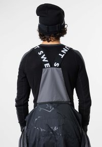 Persona che indossa una giacca nera e grigia con bretelle nere e un berretto nero, vista da dietro su uno sfondo bianco.