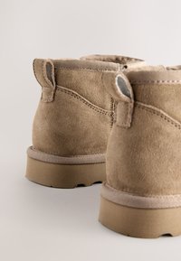 Bottes basses en daim marron clair avec une finition texturée, dotées d'un bout arrondi, d'une semelle plate et de languettes de tirage sur les côtés.