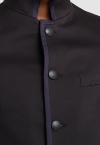 Black jacket with a high collar, featuring a contrasting purple trim, four black buttons, and a front pocket detail. Smooth fabric texture.