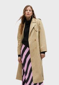 Beige double-breasted trench coat with shoulder epaulets, paired with a black turtleneck and a pink and black striped skirt.