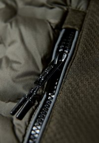 Mörk olivgrön puffjacka med detaljer som en glansig svart dragkedja, texturerat tyg och två svarta dragöglor med kontrasterande sömnad.