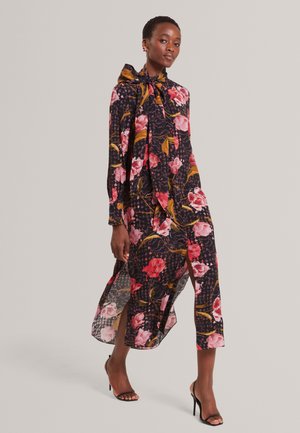 Femme portant une longue robe noire à imprimé floral rose et rouge, col montant avec nœud, fentes latérales, et sandales noires à talons hauts, marchant vers l'avant.