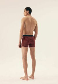 Red boxer shorts with a wave pattern, featuring a black waistband. The model is shown from the back, standing barefoot.