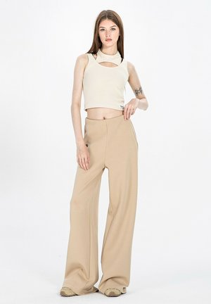 Joven mujer está de pie con pantalones beige de pierna ancha, top corto sin mangas crema con corte en el pecho y zapatillas deportivas contra un fondo blanco liso.