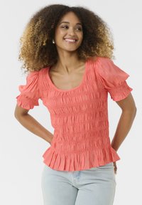 Top corallo con maniche a sbuffo, texture arricciata e orlo a volant, abbinato a jeans di denim blu chiaro. Presenta un ampio scollo a girocollo.