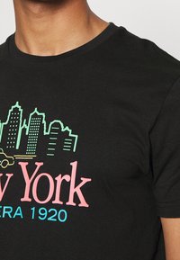 Svart bomullströja med en grafisk design av en stadsskyline i grönt och rosa, med texten "New York" och "EST. 1920".