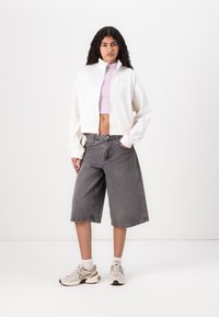 Hvid cropped jakke med høj krave, lyse lilla tætsiddende top, grå baggy shorts og beige sportssko med hvide accenter.