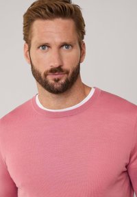 Suéter de cuello redondo rosa con una textura de punto fino, superpuesto a una camiseta blanca. El diseño presenta una silueta simple y clásica.